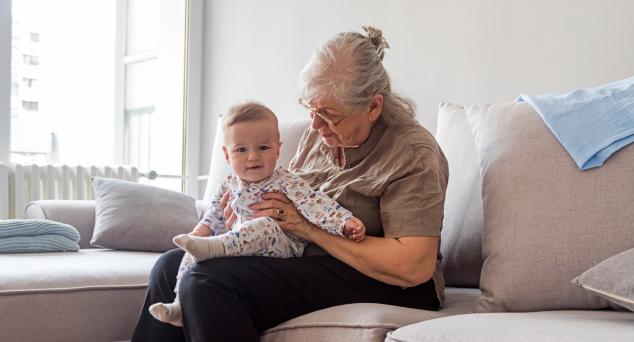 An older woman is sat with a young baby on her lap. She is looking down at the baby and the baby is smiling.