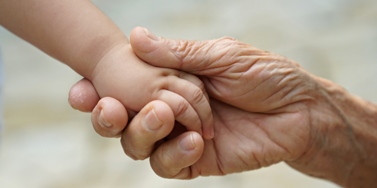 An adult hand is holding a baby's hand. Both have light skin tones.