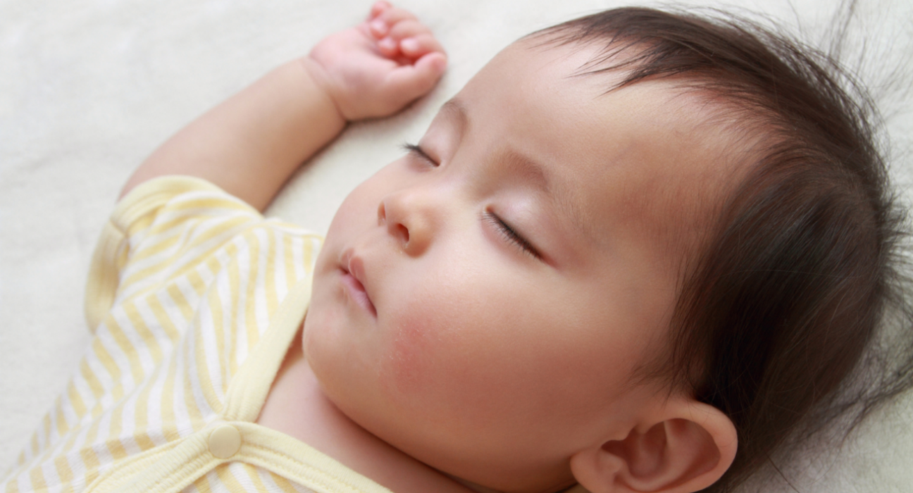 A baby is sleeping on their back. They have light skin and dark hair and wear a light yellow sleep suit.