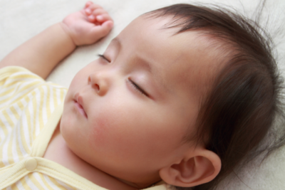 A baby is sleeping on their back. They have light skin and dark hair and wear a light yellow sleep suit.