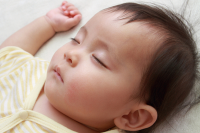 A baby is sleeping on their back. They have light skin and dark hair and wear a light yellow sleep suit.