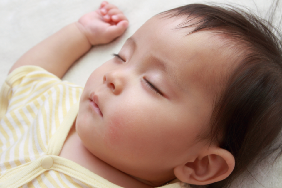 A baby is sleeping on their back. They have light skin and dark hair and wear a light yellow sleep suit.