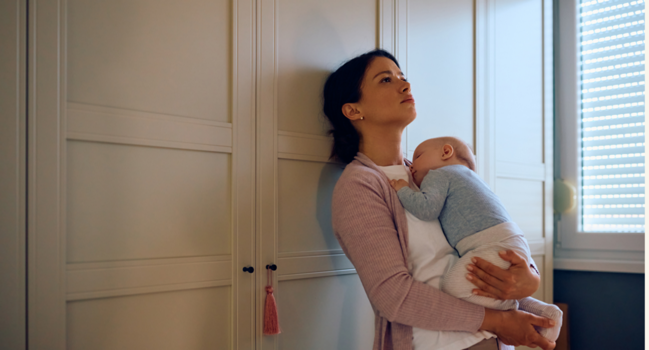 Mum is holding her baby in her arms, leaning up against a wardrobe. She looks tired and is looking at the ceiling. Her baby is asleep on her chest.