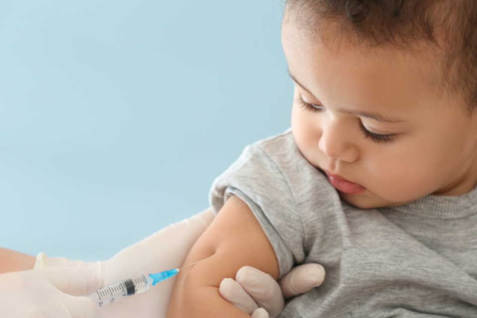 A baby is receiving a vaccination. They are looking down at the needle curiously. They have olive skin and dark hair.