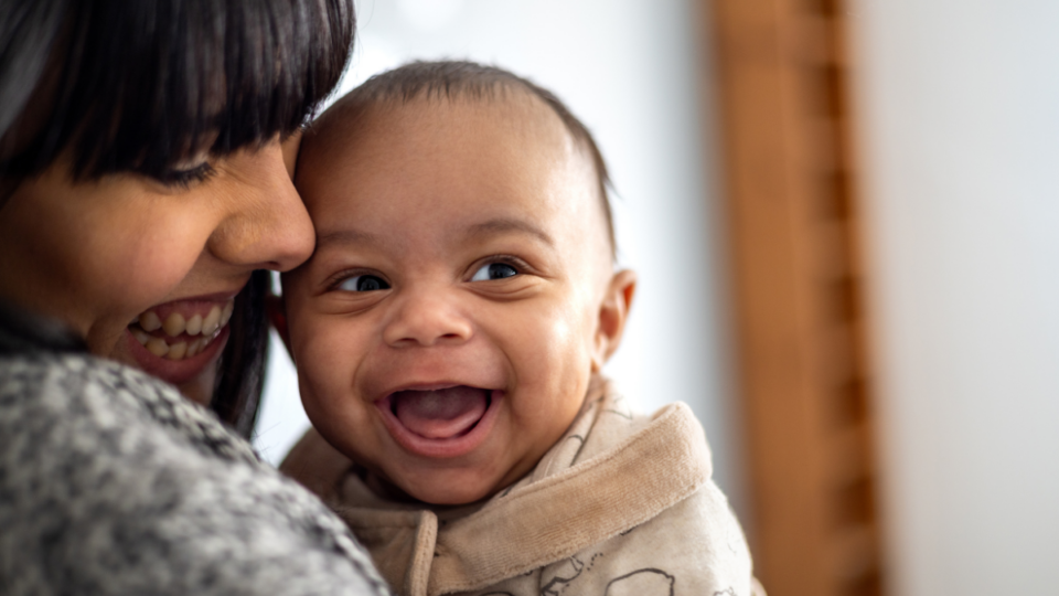 A baby is looking to their side with a huge, wide open grin. They have light to medium skin and the mum is looking down at them and smiling, with her face closely pressed to the baby.