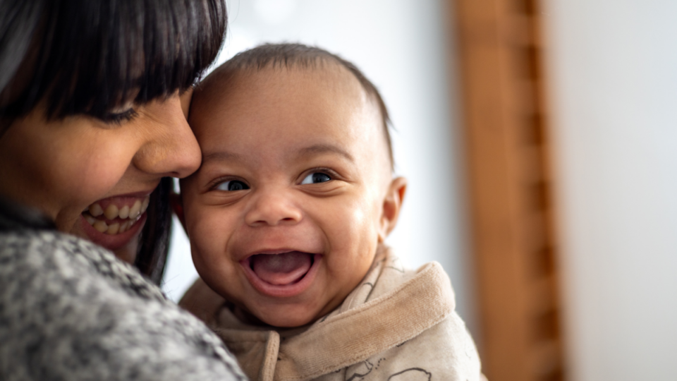 A baby is looking to their side with a huge, wide open grin. They have light to medium skin and the mum is looking down at them and smiling, with her face closely pressed to the baby.