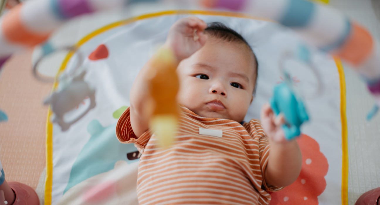 A baby is laying on their back playing with toys dangling above. They are wearing an orange and white stripey baby grow.