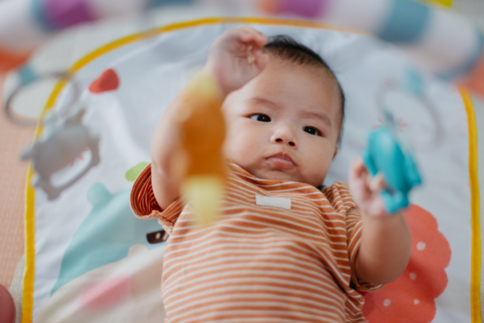 A baby is laying on their back playing with toys dangling above. They are wearing an orange and white stripey baby grow.