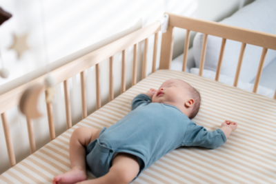 A baby in a blue baby grow is sleeping on their back in a wooden cot with a firm mattress.