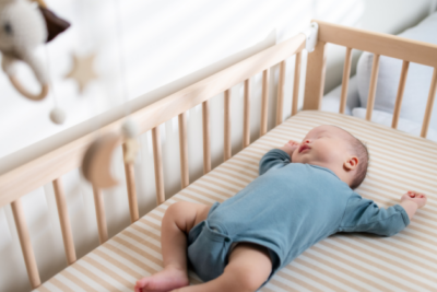 A baby in a blue baby grow is sleeping on their back in a wooden cot with a firm mattress.