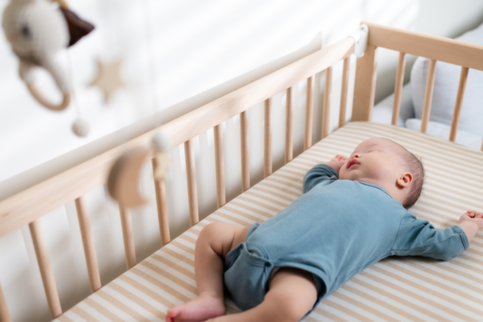A baby in a blue baby grow is sleeping on their back in a wooden cot with a firm mattress.
