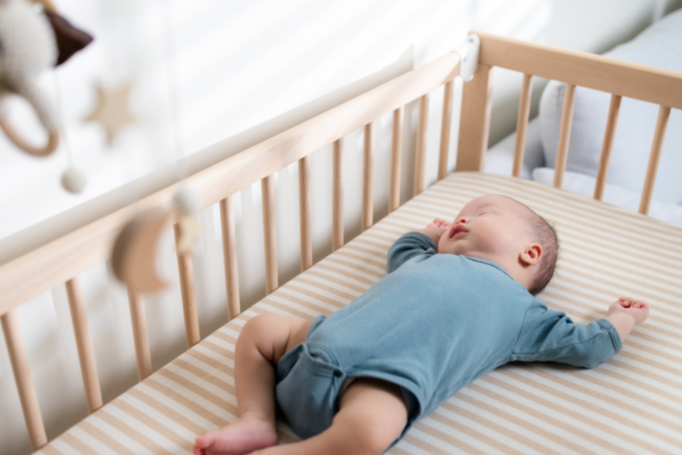 A baby in a blue baby grow is sleeping on their back in a wooden cot with a firm mattress.