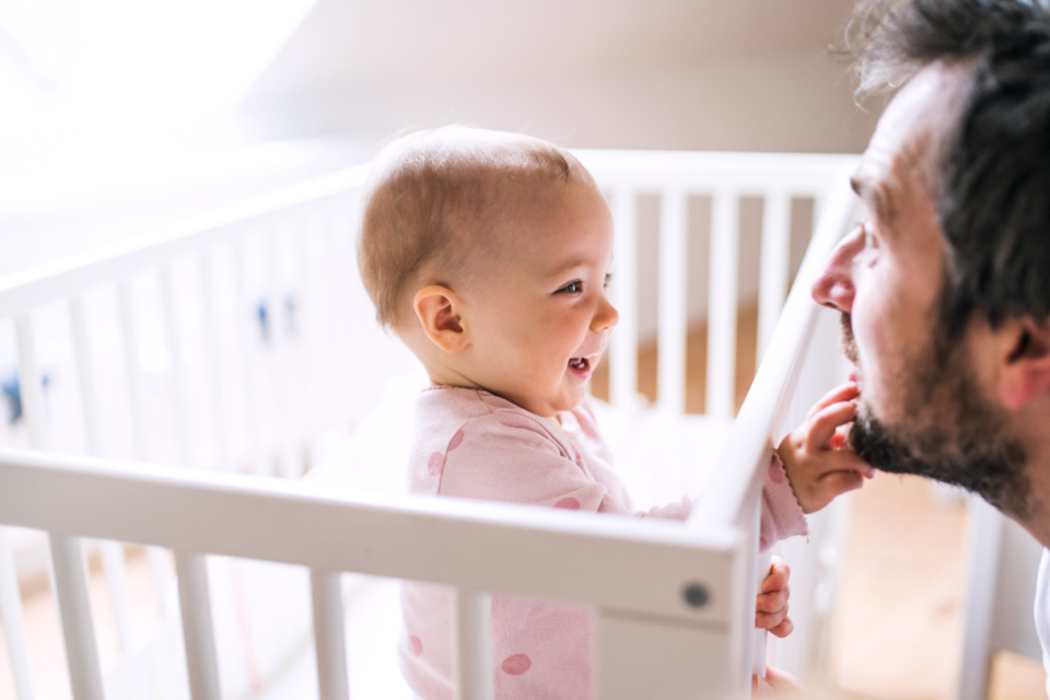 A dad is at face-level with a baby in a cot. The baby is smiling and playing with his beard.