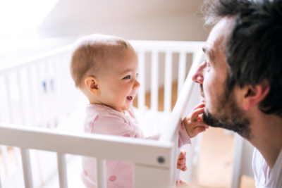 A dad is at face-level with a baby in a cot. The baby is smiling and playing with his beard.