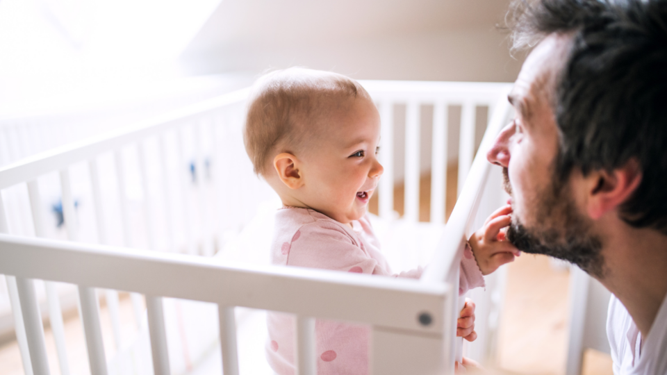 A dad is at face-level with a baby in a cot. The baby is smiling and playing with his beard.