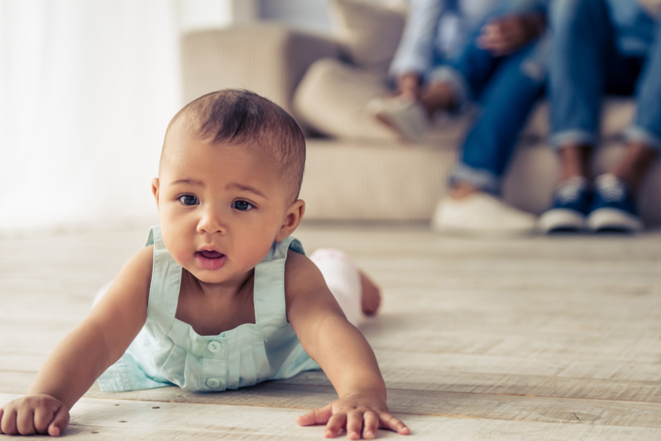 Baby is spending time on their tummy in the living room. They have brown skin and hair, and wearing a blue vest dress.