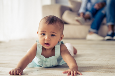 Baby is spending time on their tummy in the living room. They have brown skin and hair, and wearing a blue vest dress.