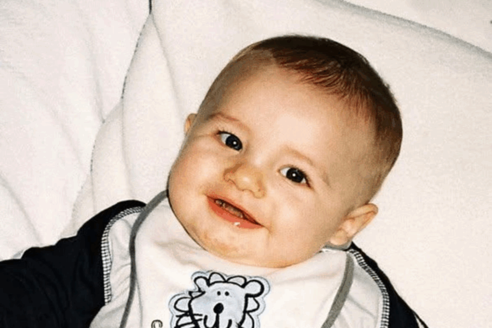 Harvey is sat against something cushiony. He has very sharp eyes, a small nose and plump cheeks. He has fair skin, light brown hair and is smiling, wearing a bib.