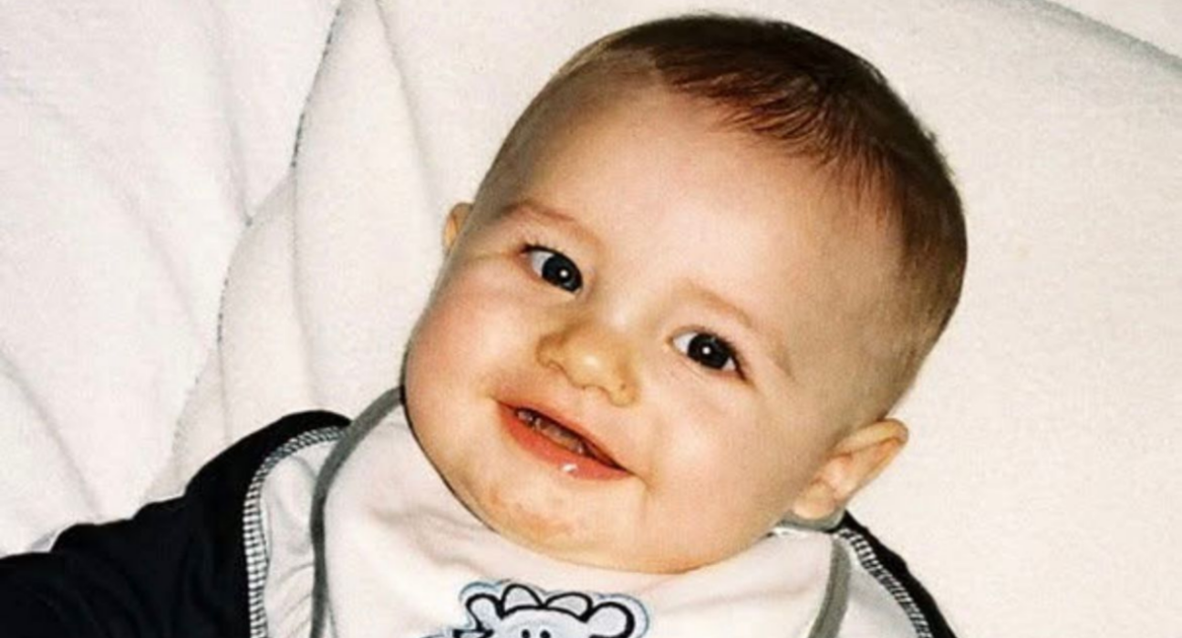 Harvey is sat against something cushiony. He has very sharp eyes, a small nose and plump cheeks. He has fair skin, light brown hair and is smiling, wearing a bib.