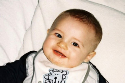 Harvey is sat against something cushiony. He has very sharp eyes, a small nose and plump cheeks. He has fair skin, light brown hair and is smiling, wearing a bib.