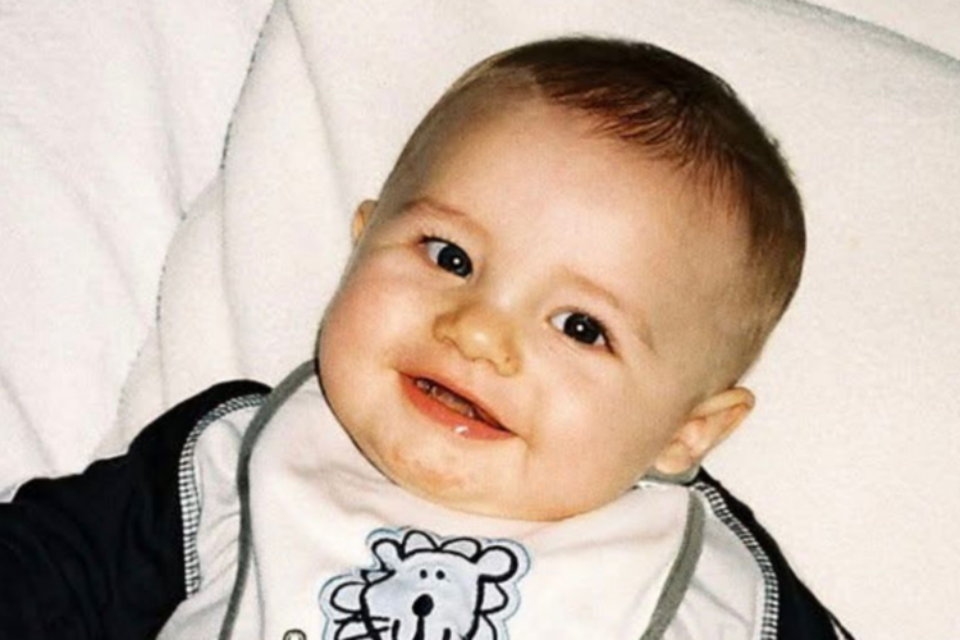 Harvey is sat against something cushiony. He has very sharp eyes, a small nose and plump cheeks. He has fair skin, light brown hair and is smiling, wearing a bib.