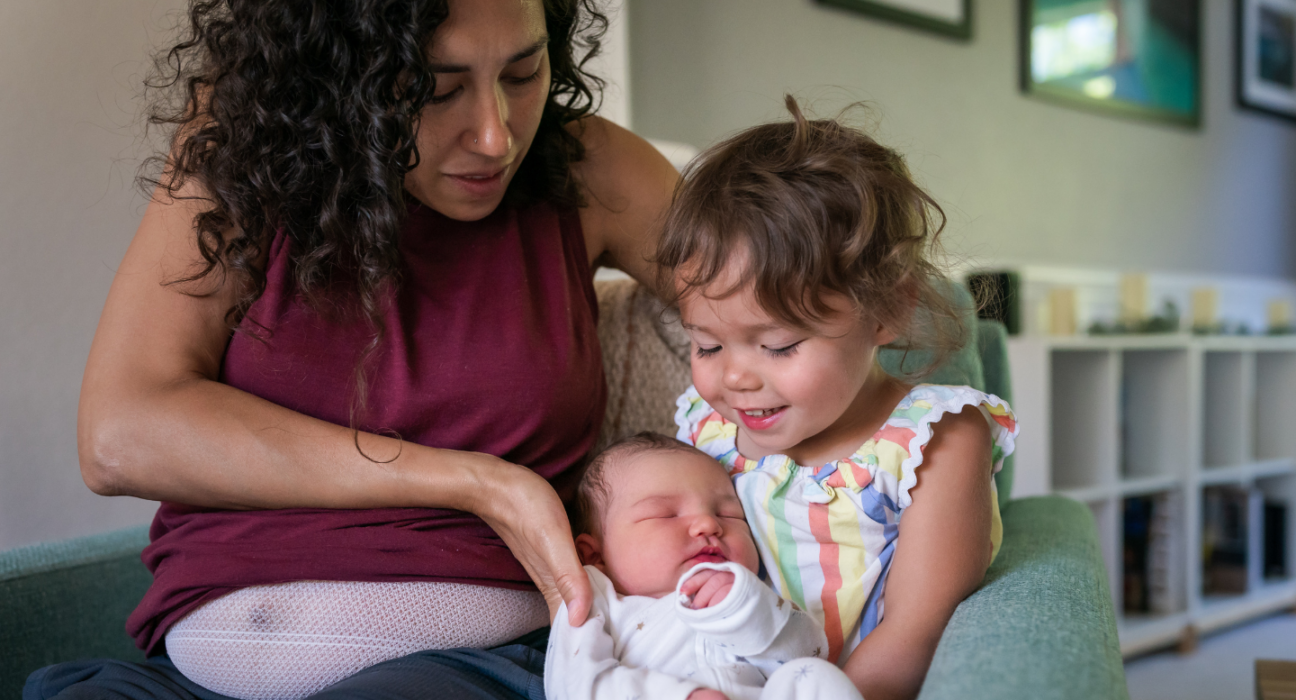 A mum is placing her new born in the arms of the older sibling. They are sat together on a couch.