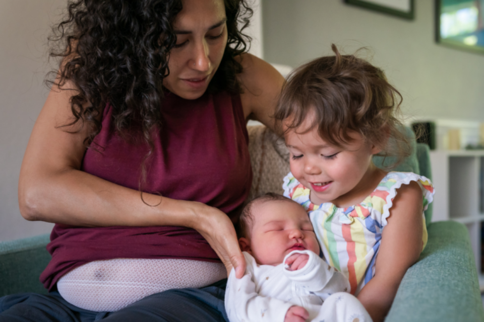 A mum is placing her new born in the arms of the older sibling. They are sat together on a couch.
