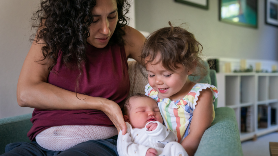 A mum is placing her new born in the arms of the older sibling. They are sat together on a couch.