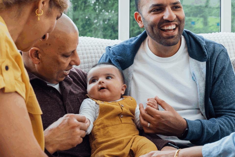 A family are gathered around their baby. They are all smiling and looking at the baby and the baby is sat in between two of the men.