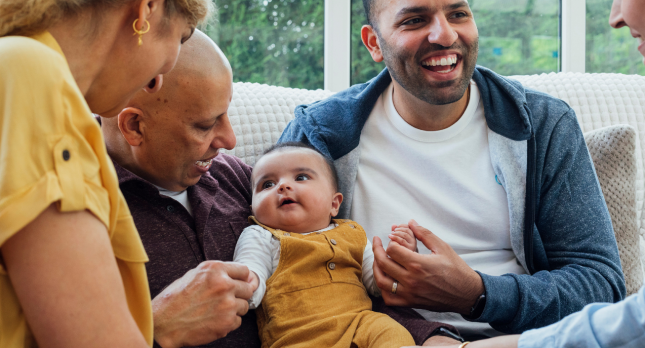 A family are gathered around their baby. They are all smiling and looking at the baby and the baby is sat in between two of the men.