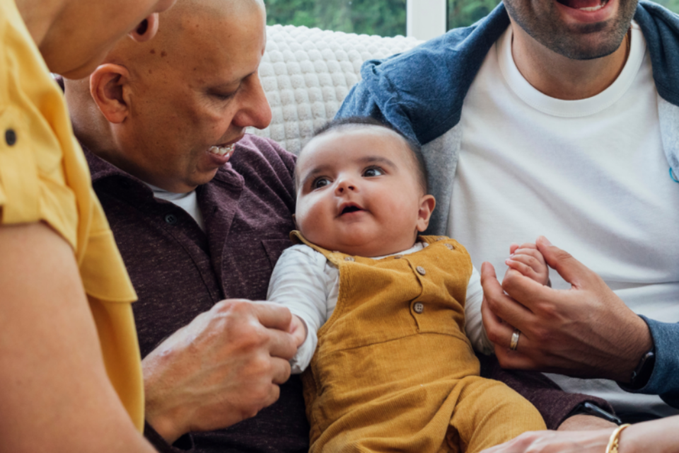 A family are gathered around their baby. They are all smiling and looking at the baby and the baby is sat in between two of the men.
