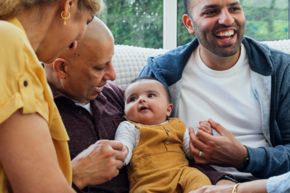 A family are gathered around their baby. They are all smiling and looking at the baby and the baby is sat in between two of the men.