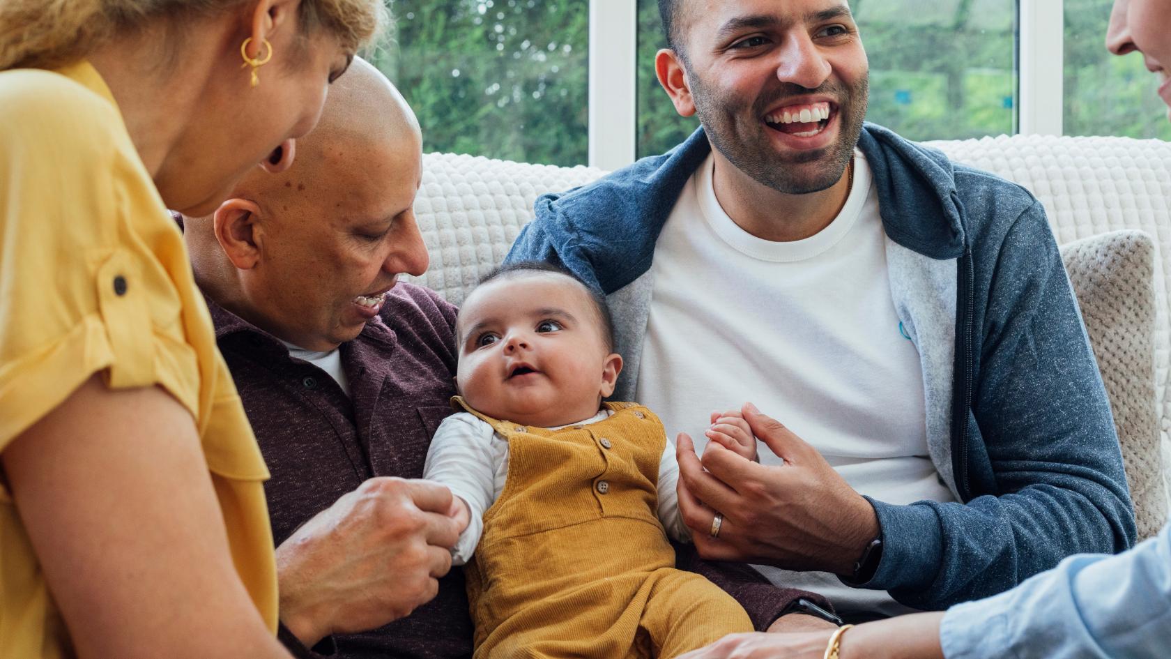A family are gathered around their baby. They are all smiling and looking at the baby and the baby is sat in between two of the men.