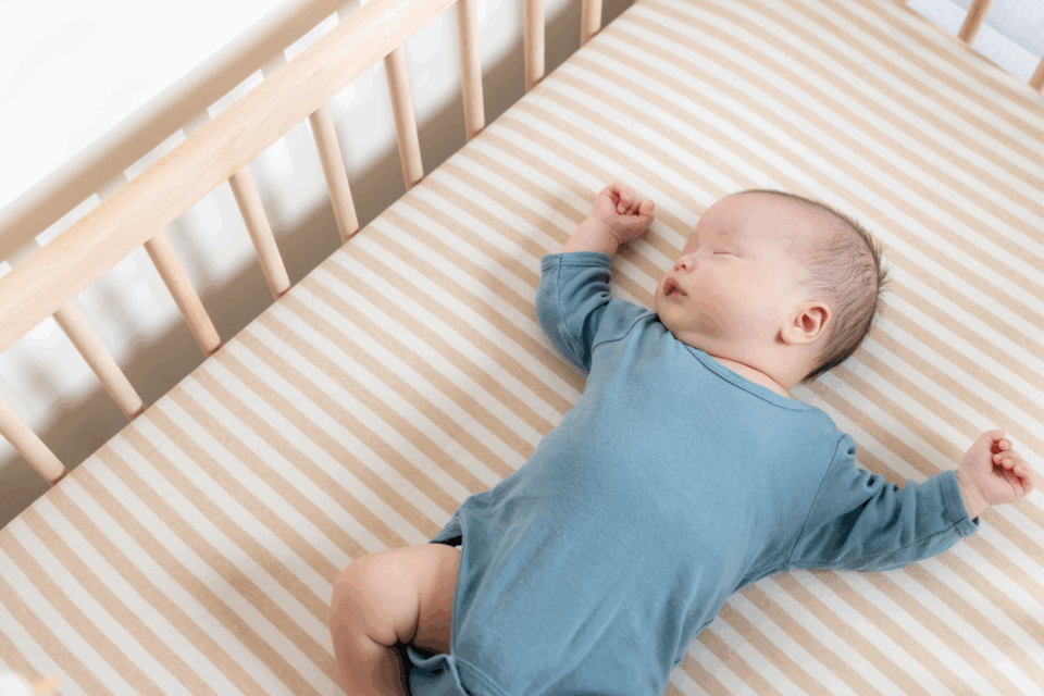 A baby in a blue baby grow is sleeping on their back in a wooden cot with a firm mattress.