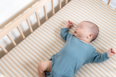 A baby in a blue baby grow is sleeping on their back in a wooden cot with a firm mattress.