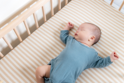 A baby in a blue baby grow is sleeping on their back in a wooden cot with a firm mattress.