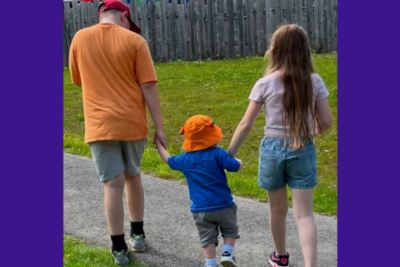 Bailey Poppy and Jack are walking with their backs to the camera. The eldest are holding the hands of the little one, Jack's, hand in the middle. They are dressed in summer clothes.