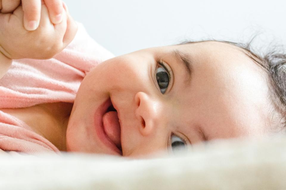 A close up of a baby on their side, smiling. Their tongue is slightly sticking out, they have light skin and dark curly hair and their hands are clasped just in view.