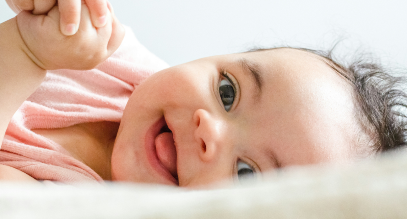 A close up of a baby on their side, smiling. Their tongue is slightly sticking out, they have light skin and dark curly hair and their hands are clasped just in view.