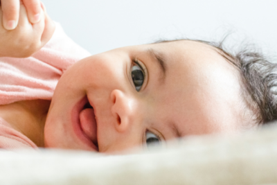 A close up of a baby on their side, smiling. Their tongue is slightly sticking out, they have light skin and dark curly hair and their hands are clasped just in view.