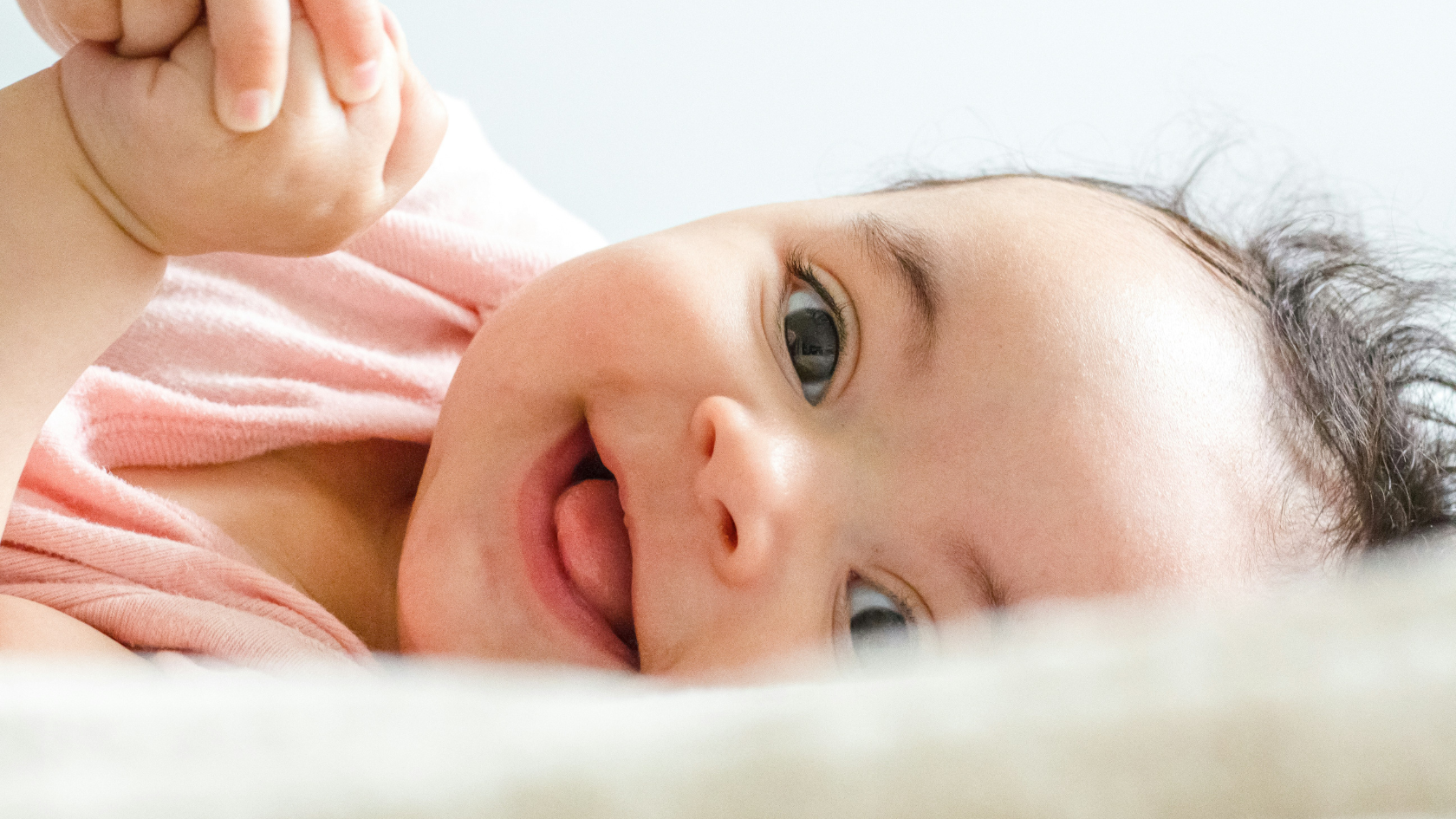A close up of a baby on their side, smiling. Their tongue is slightly sticking out, they have light skin and dark curly hair and their hands are clasped just in view.