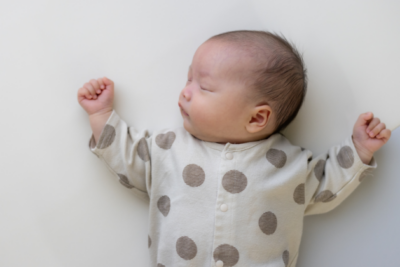 A baby is sleeping in on their back, with their head tilted to the side. They have dark hair, light skin and wear a polkadot baby grow. Their arms are rested either side of their head.