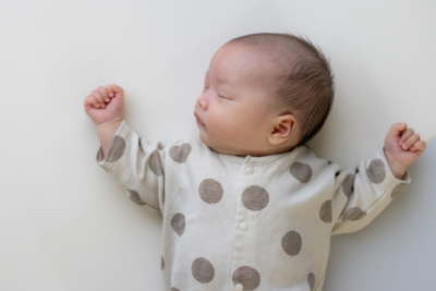 A baby is sleeping in on their back, with their head tilted to the side. They have dark hair, light skin and wear a polkadot baby grow. Their arms are rested either side of their head.
