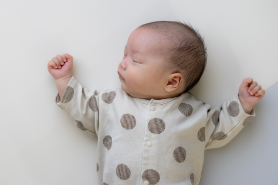 A baby is sleeping in on their back, with their head tilted to the side. They have dark hair, light skin and wear a polkadot baby grow. Their arms are rested either side of their head.