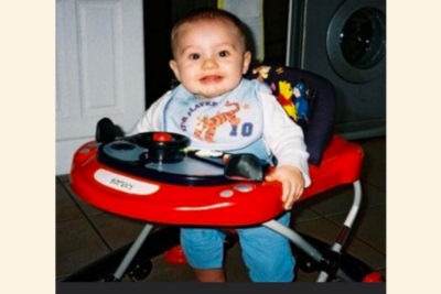 Baby Harvey is sat in a baby walker. He has a very wide and warm grin. He has fair skin and light brown hair.