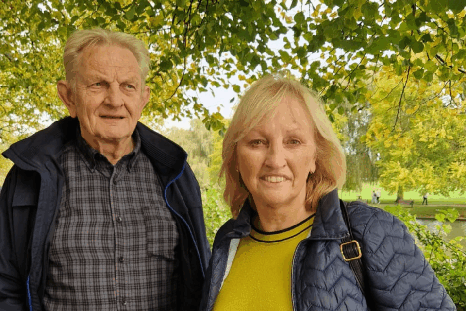 Bridget and Zbig are in a park surrounded by greenery. They are both smiling. Bridget is looking straight on, and Zbig to the side.