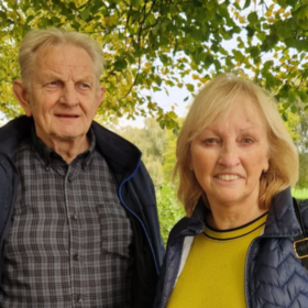 Bridget and Zbig are in a park surrounded by greenery. They are both smiling. Bridget is looking straight on, and Zbig to the side.