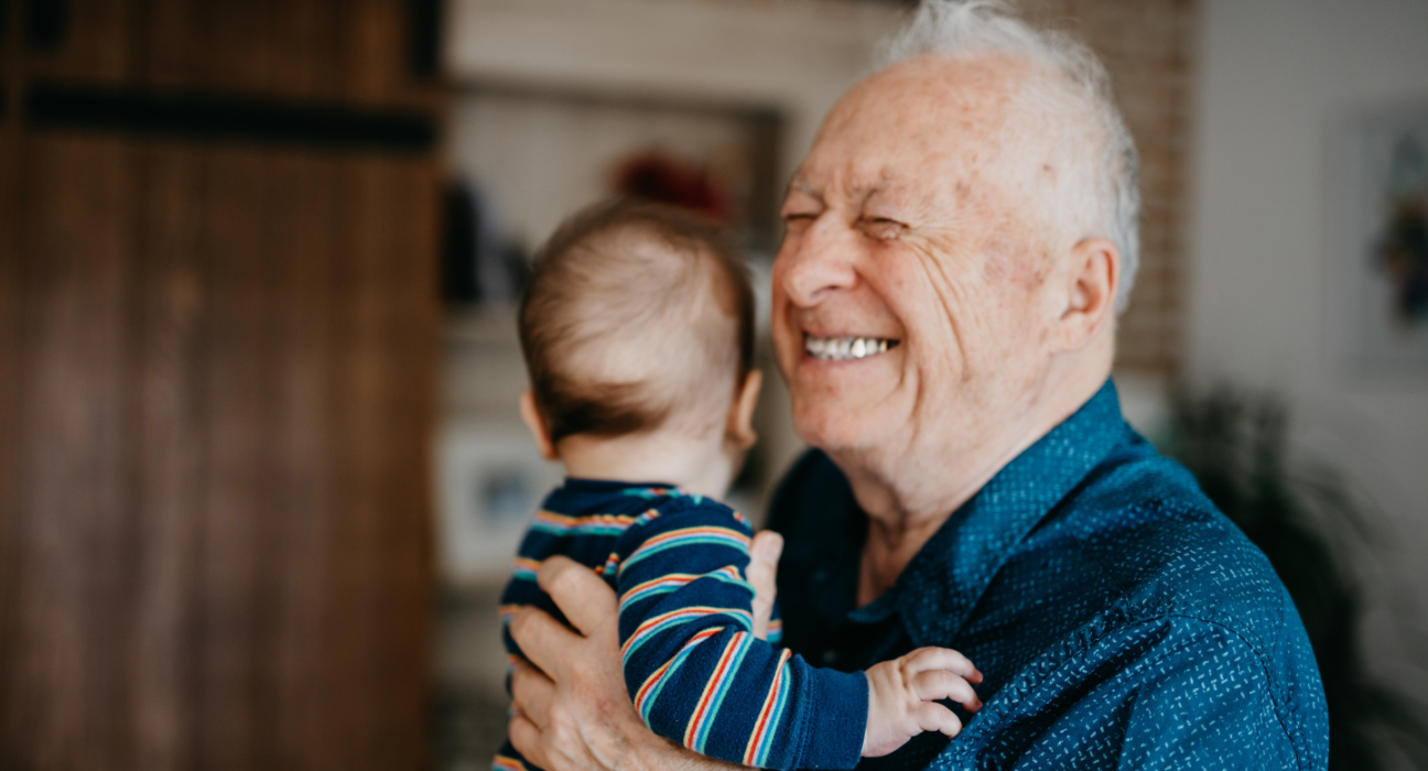 Grandad standing up smiling, holding a baby who is looking away.