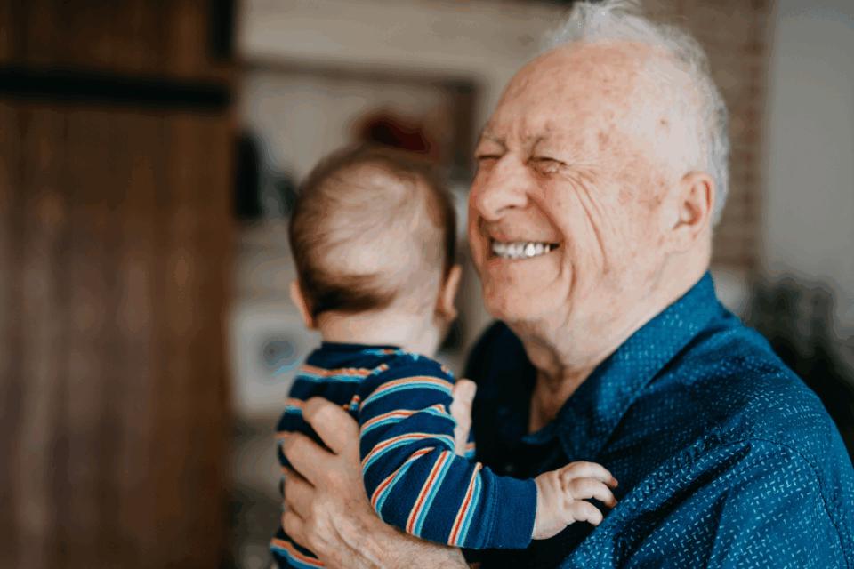 Grandad standing up smiling, holding a baby who is looking away.