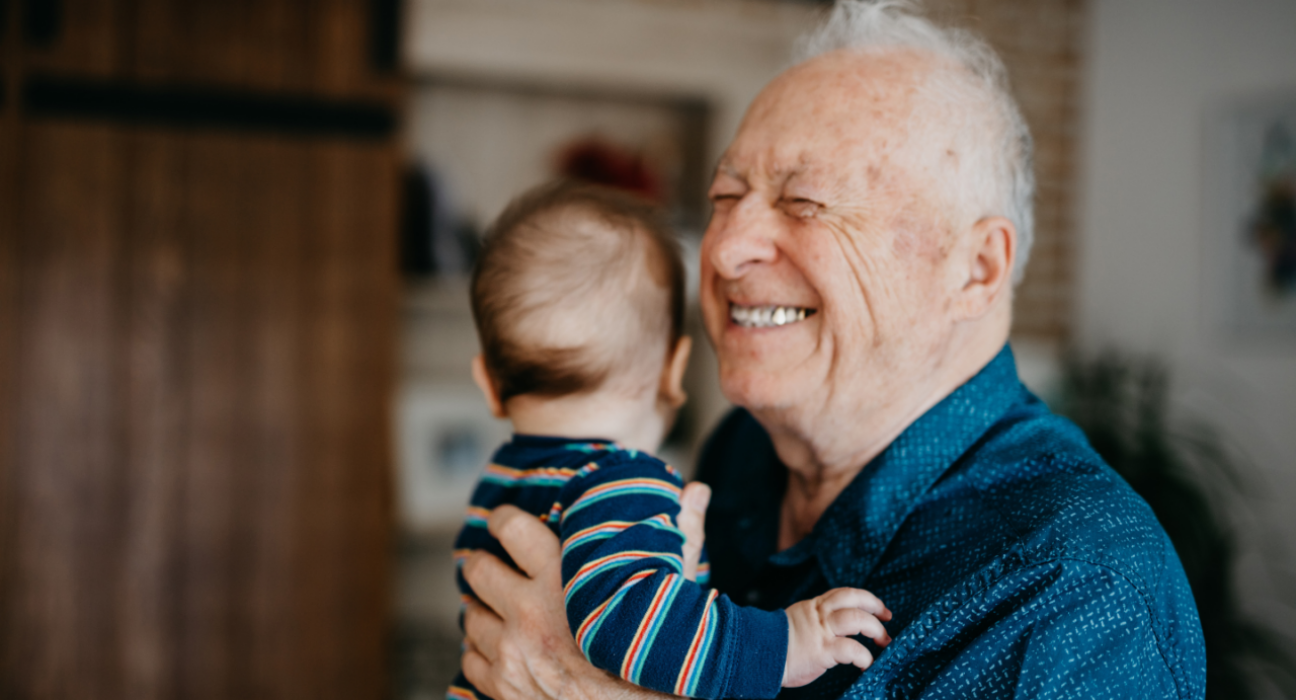 Grandad standing up smiling, holding a baby who is looking away.